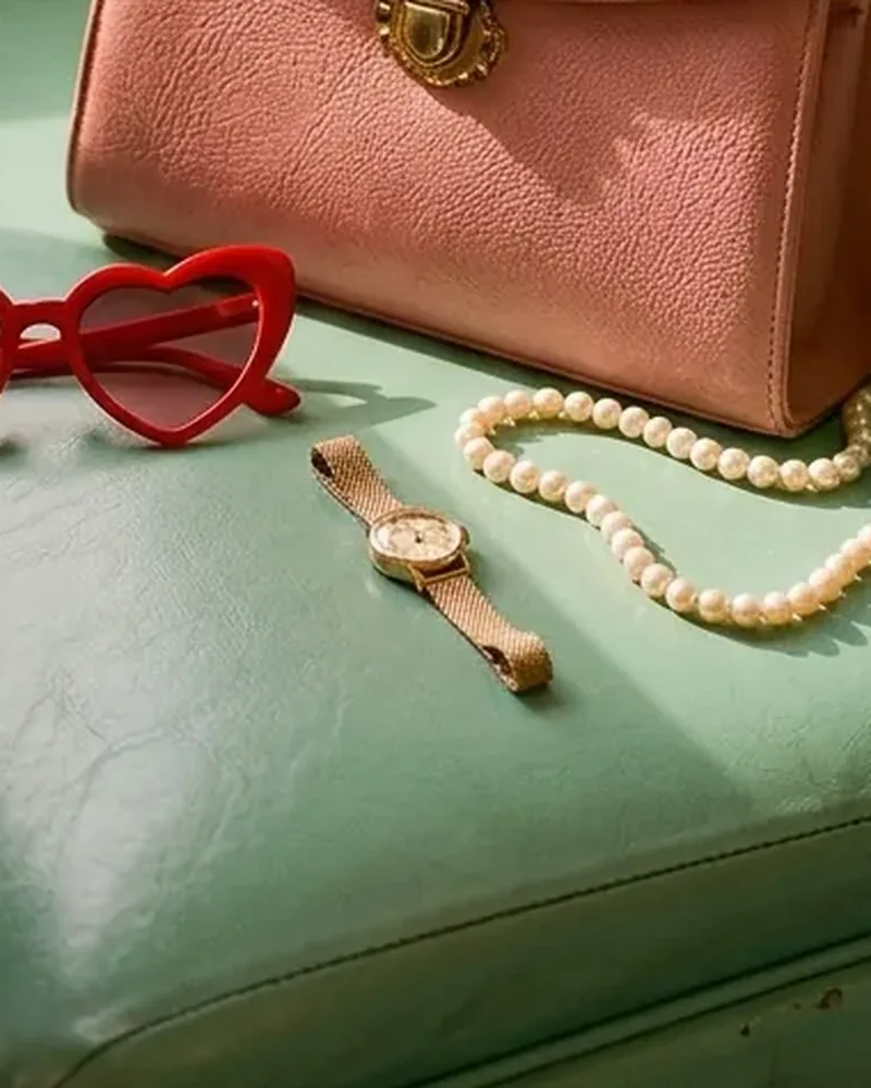 Reloj dorado de malla junto a unas gafas y un collar de perlas